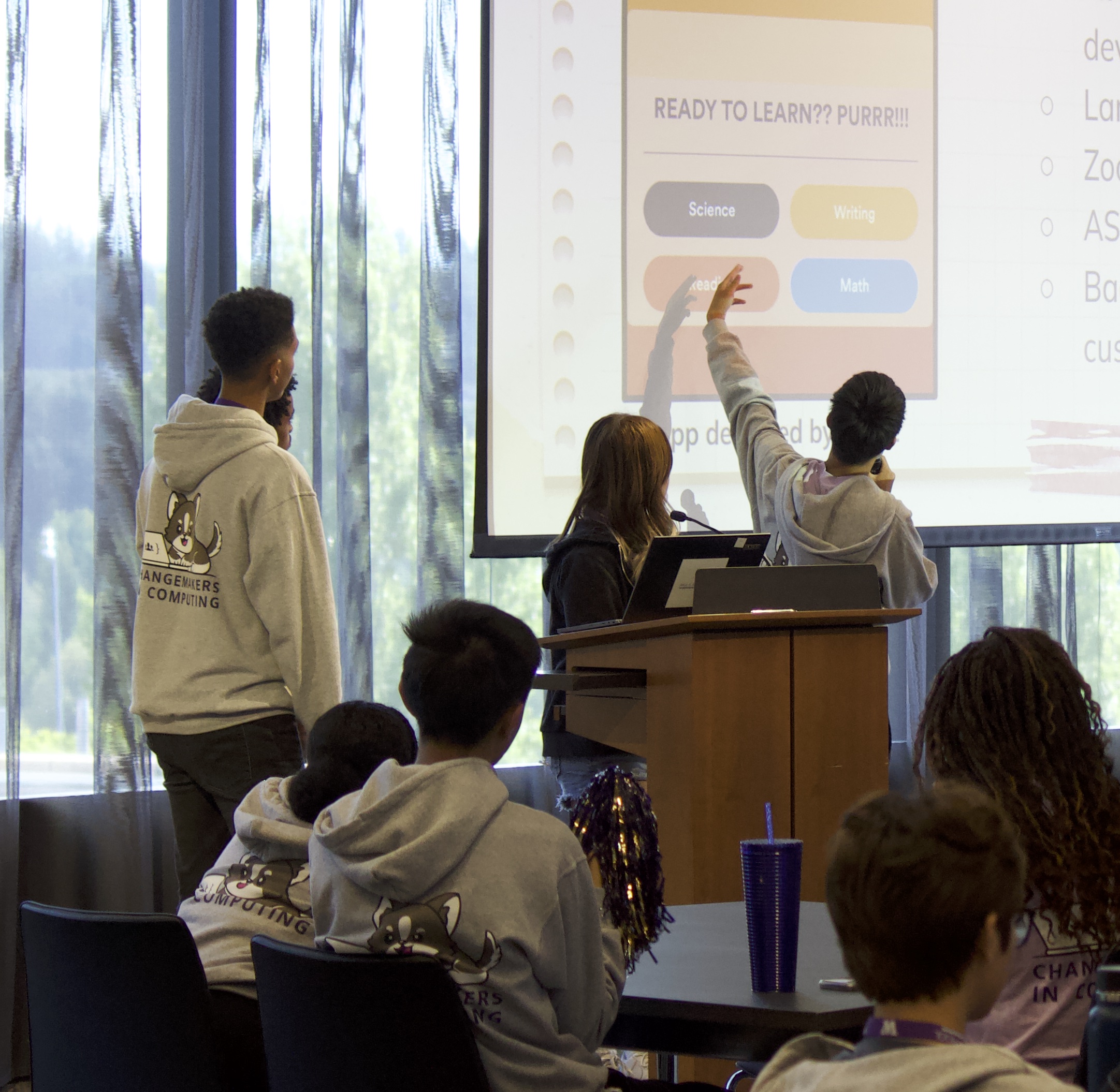 A student with a microphone gesturing towards a screen with students sitting nearby watching